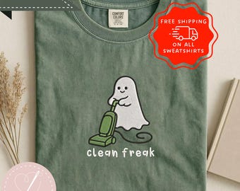 May include: A moss green crew neck t-shirt featuring an embroidered graphic of a white ghost using a green vacuum cleaner. Below the ghost, the text reads clean freak in white lowercase letters. The shirt has a Comfort Colors brand label.