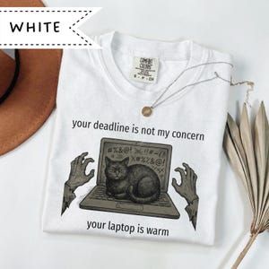 Pode incluir: Camiseta branca com o texto "your deadline is not my concern" e "your laptop is warm". O gráfico mostra um gato preto em um laptop com mãos de zumbi. Um colar de prata com um pingente também é visível.