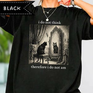 Puede incluir: Una camiseta negra con un estampado gráfico de un gato negro sentado en un taburete frente a un espejo. El texto sobre la imagen dice i do not think y el texto debajo dice therefore i do not am. La ilustración tiene un estilo de boceto.