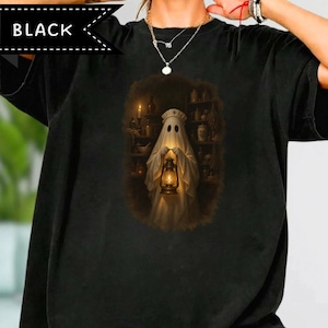May include: A black t-shirt featuring a graphic print of a ghost dressed as a nurse, wearing a cap and holding a glowing lantern. The ghost is positioned in front of shelves filled with apothecary jars and bottles in a dimly lit, dark setting.