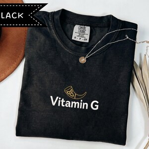 May include: Black t-shirt with the text "Vitamin G" and a gold-colored graphic. The shirt has a crew neck and short sleeves. A silver necklace with a round pendant is on the shirt. The shirt is labeled "Comfort Colors".