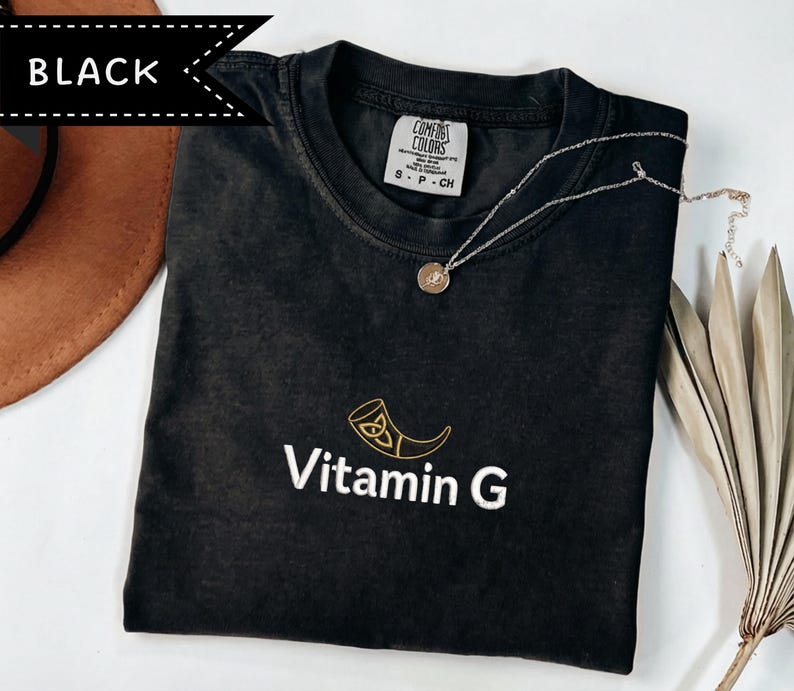 May include: Black t-shirt with the text "Vitamin G" and a gold-colored graphic. The shirt has a crew neck and short sleeves. A silver necklace with a round pendant is on the shirt. The shirt is labeled "Comfort Colors".