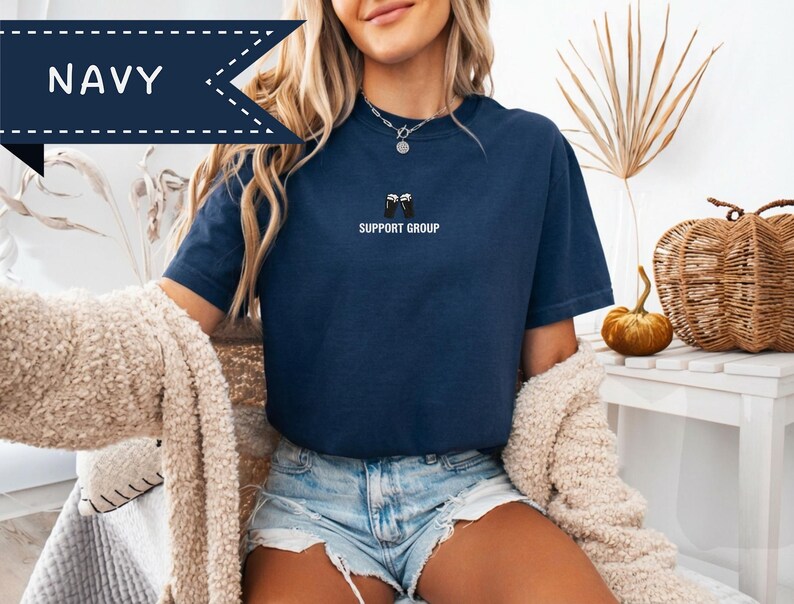 May include: Navy blue t-shirt with the text "SUPPORT GROUP" in white. The shirt has a small graphic of two black mugs. The word "NAVY" is displayed in a navy blue banner. The model is wearing denim shorts and a cream-colored sweater.