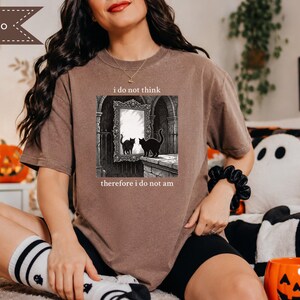 Puede incluir: Una camiseta color espresso con un estampado gráfico de un gato negro de pie sobre una repisa de piedra mirando hacia un espejo ornamentado. Sobre el espejo está el texto i do not think y debajo el texto therefore i do not am.