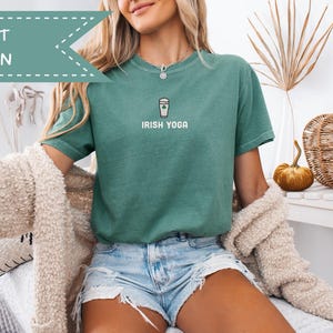 May include: A light green t-shirt with the text "IRISH YOGA" and a graphic of a coffee cup with a shamrock. The shirt is paired with denim shorts. A light-colored sweater is draped over the shoulders.
