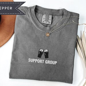 May include: A gray t-shirt with the words "SUPPORT GROUP" printed below a graphic of two beer glasses. A gold necklace with a pendant rests on the shirt. The shirt is folded and placed on a white surface.