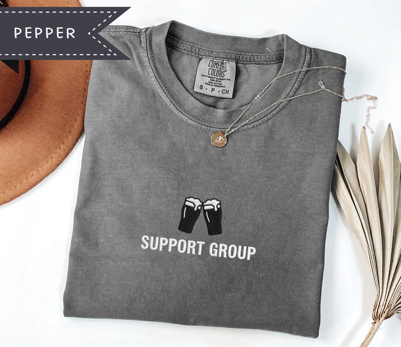 May include: A gray t-shirt with the words "SUPPORT GROUP" printed below a graphic of two beer glasses. A gold necklace with a pendant rests on the shirt. The shirt is folded and placed on a white surface.
