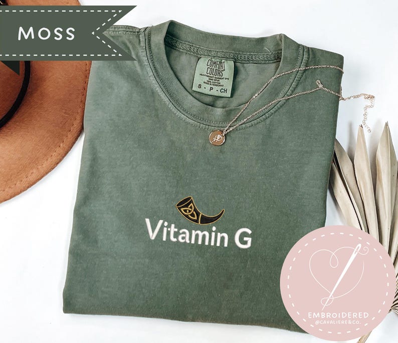May include: A moss green t-shirt with the embroidered text "Vitamin G" and a horn graphic. A gold necklace with a pendant rests on the shirt. The shirt is a Comfort Colors brand.
