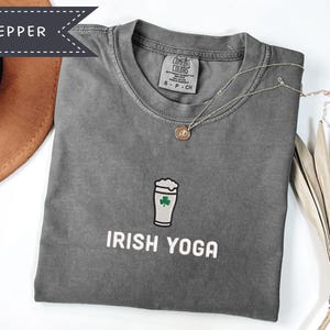 May include: A gray t-shirt with the words "IRISH YOGA" printed in white. The shirt features a graphic of a beer glass with a shamrock. A gold necklace with a pendant is on the shirt. The shirt is folded on a white surface.