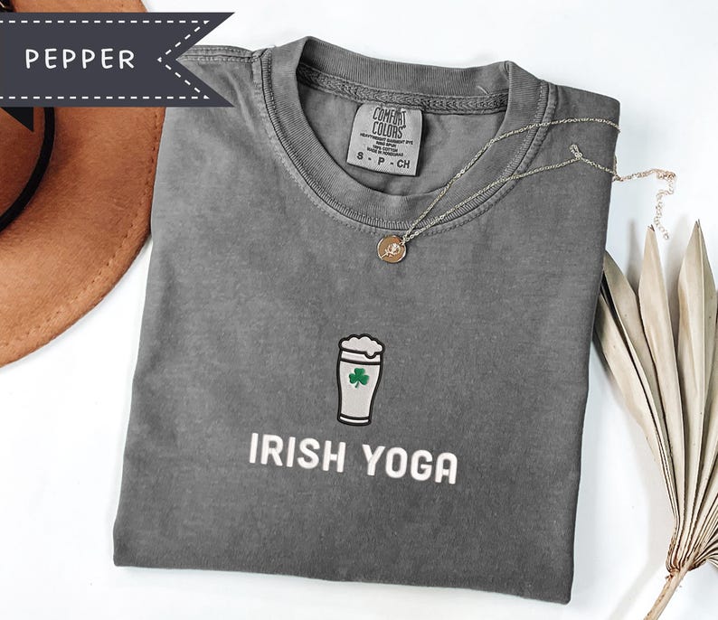 May include: A gray t-shirt with the words "IRISH YOGA" printed in white. The shirt features a graphic of a beer glass with a shamrock. A gold necklace with a pendant is on the shirt. The shirt is folded on a white surface.