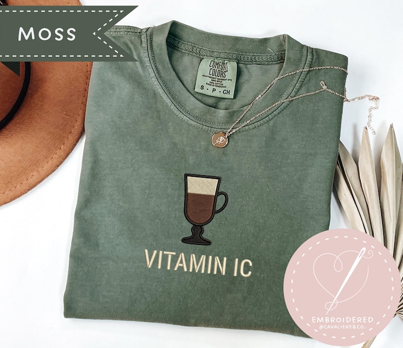 May include: A moss green t-shirt with an embroidered coffee drink design and the text "VITAMIN IC". The shirt has a crew neck and short sleeves. A gold necklace is draped across the shirt. The shirt is part of a clothing collection.