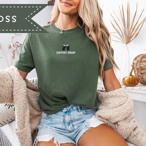 May include: A moss green t-shirt with the text "SUPPORT GROUP" and a graphic of two beer mugs. The shirt is paired with denim shorts. The word "MOSS" is in a banner in the upper left corner.