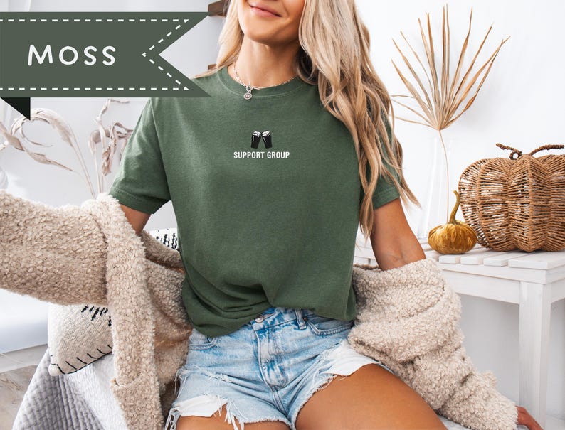May include: A moss green t-shirt with the text "SUPPORT GROUP" and a graphic of two beer mugs. The shirt is paired with denim shorts. The word "MOSS" is in a banner in the upper left corner.