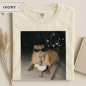 May include: An ivory t-shirt featuring a graphic print of a brown capybara wearing black sunglasses and drinking from a glass with a lime wedge and a straw. The shirt has a crew neckline and a Comfort Colors brand label on the inside collar.