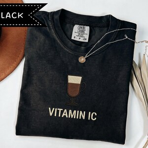 May include: Black t-shirt with an embroidered coffee drink design and the text "VITAMIN IC" in gold. A silver necklace with a small pendant rests on the shirt. The shirt is a Comfort Colors brand.