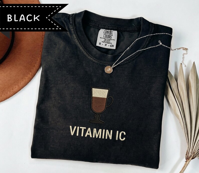 May include: Black t-shirt with an embroidered coffee drink design and the text "VITAMIN IC" in gold. A silver necklace with a small pendant rests on the shirt. The shirt is a Comfort Colors brand.