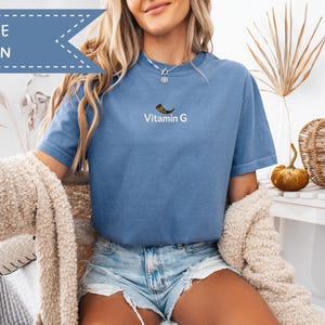 May include: Blue jean t-shirt with the text "Vitamin G" and a graphic of a horn. The shirt is a light blue color. A banner that says "Blue Jean" is in the upper left corner.