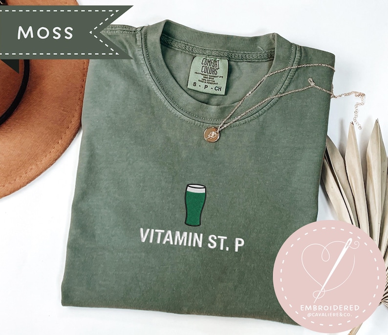 May include: A moss green t-shirt with a green beer glass graphic and the text "Vitamin St. P". The shirt has a crew neck and short sleeves. A gold necklace is draped across the shirt. A brown hat is in the background.