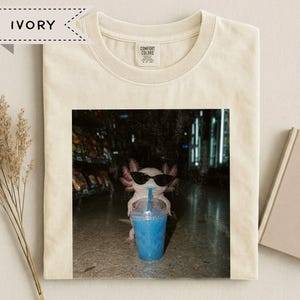 Könnte beinhalten: Ein elfenbeinfarbenes T-Shirt mit einem Grafikdruck eines Axolotls, das eine schwarze Sonnenbrille trägt und einen durchsichtigen Plastikbecher mit einem leuchtend blauen Getränk hält. Das Shirt hat einen Rundhalsausschnitt und ein Comfort Colors Etikett.