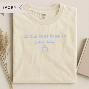 May include: An ivory short-sleeve t-shirt with a crew neckline. The center of the shirt features the light blue text "on the same team as your kid" positioned above a simple line-drawn apple icon. The interior neck label reads Comfort Colors.