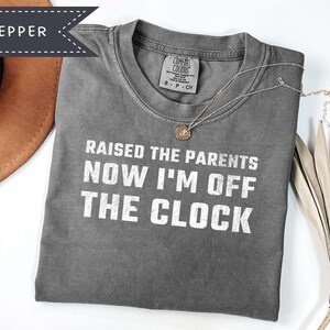 May include: A pepper-colored short-sleeve t-shirt featuring the white text RAISED THE PARENTS NOW I'M OFF THE CLOCK printed on the front. The shirt has a crew neckline and a Comfort Colors brand label on the inside collar area.