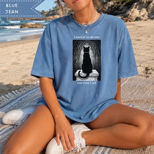 May include: A blue jean colored short-sleeve t-shirt featuring a graphic print of a black cat sitting on a stone pedestal in front of a gothic archway. The text above the cat reads i answer to no one and below the cat reads not even God.