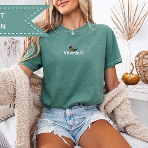 May include: Light green t-shirt with the text "Vitamin G" and a graphic. The shirt is a solid color and has short sleeves. The image also shows denim shorts and a cream-colored cardigan.