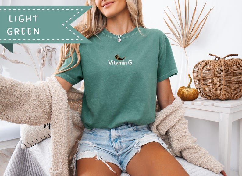 May include: Light green t-shirt with the text "Vitamin G" and a graphic. The shirt is a solid color and has short sleeves. The image also shows denim shorts and a cream-colored cardigan.