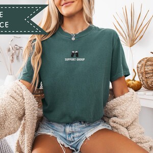 May include: Blue spruce t-shirt with the text "SUPPORT GROUP" and a graphic of two beer mugs clinking. The shirt is a solid teal color. The words "BLUE SPRUCE" are in a banner at the top left.