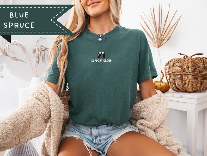 May include: Blue spruce t-shirt with the text "SUPPORT GROUP" and a graphic of two beer mugs clinking. The shirt is a solid teal color. The words "BLUE SPRUCE" are in a banner at the top left.