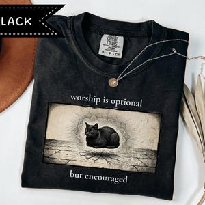 Może przedstawiać: Czarny t-shirt z grafiką przedstawiającą czarnego kota siedzącego na popękanej powierzchni z motywem mandali w tle. Tekst nad grafiką brzmi "worship is optional", a tekst pod grafiką brzmi "but encouraged".