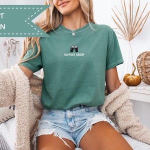 May include: Light green t-shirt with the text "SUPPORT GROUP" and a graphic of two beer mugs. The shirt is paired with denim shorts. The image also includes a beige cardigan, a decorative pumpkin, and a woven basket.