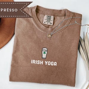 May include: Espresso-colored t-shirt with the words "IRISH YOGA" printed in white. The shirt features a graphic of a beer glass with a green shamrock. A silver necklace with a pendant is draped across the shirt.