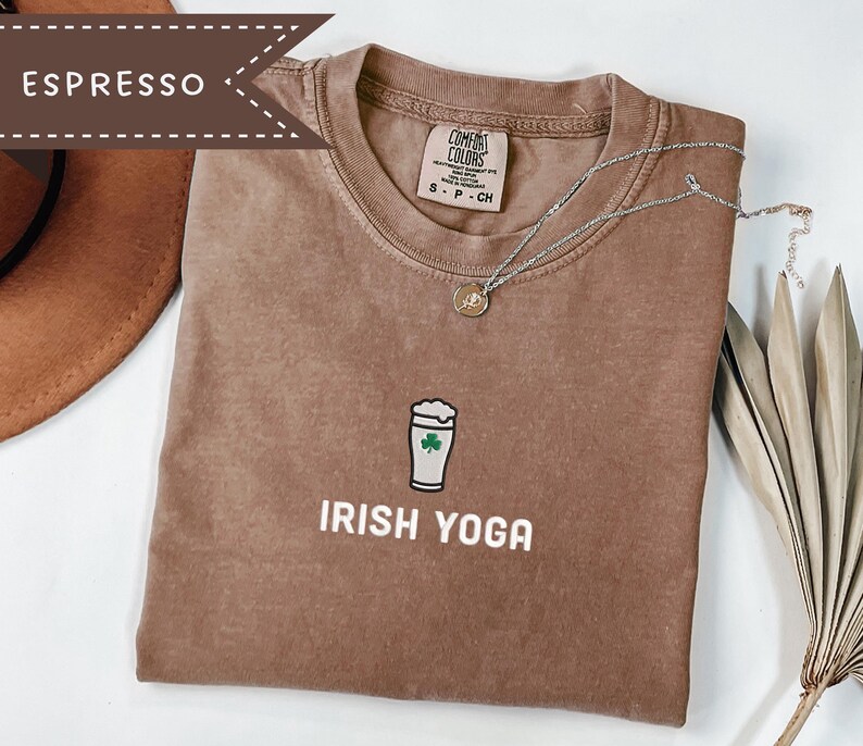 May include: Espresso-colored t-shirt with the words "IRISH YOGA" printed in white. The shirt features a graphic of a beer glass with a green shamrock. A silver necklace with a pendant is draped across the shirt.