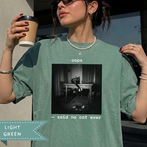 Puede incluir: Una camiseta verde claro con un gráfico en blanco y negro de un gato sobre una mesa tirando un objeto. El texto sobre el gráfico dice "oops" y el texto debajo dice "- said no cat ever". La camiseta tiene cuello redondo y mangas cortas.