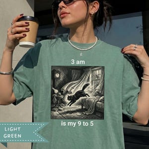 Puede incluir: Una camiseta verde claro con un estampado gráfico de un gato negro saltando por un dormitorio de noche. El texto sobre la imagen dice 3 am y el texto debajo dice is my 9 to 5. La ilustración incluye un reloj de pie, cortinas y una cama.