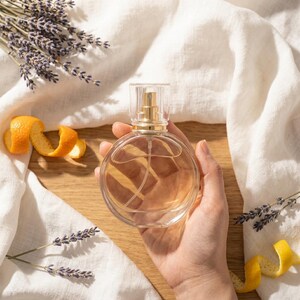 May include: A round, clear glass perfume bottle with a gold-toned spray nozzle, filled with a light amber liquid. The bottle is surrounded by sprigs of dried purple lavender and curled orange peels on a light-colored fabric background.