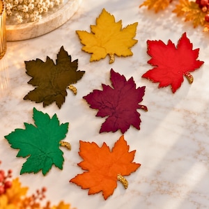 May include: A set of six maple leaf-shaped coasters in yellow, red, dark brown, purple, green, and orange. Each coaster features a textured surface and a small, gold-toned glittery stem detail at the base of the leaf, arranged in a cluster.
