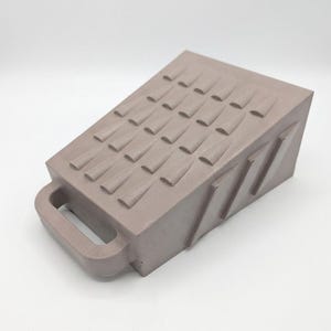 May include: A taupe-colored wedge-shaped doorstop featuring a handle on the front and a textured top surface with rows of raised, triangular protrusions. The side of the object displays three diagonal recessed grooves, providing a functional design.