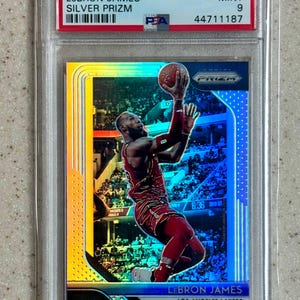 Puede incluir: Una tarjeta de baloncesto Panini Prizm LeBron James Silver Prizm de 2018, calificada como Mint 9 por PSA. La tarjeta presenta a LeBron James con un uniforme rojo de Los Angeles Lakers, en acción con un balón de baloncesto. La tarjeta está en un soporte protector.