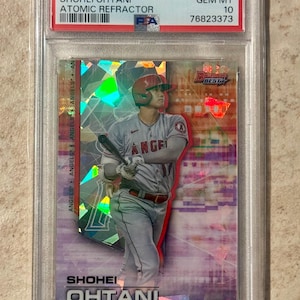 May include: A 2021 Bowman's Best Shohei Ohtani Atomic Refractor baseball card, graded GEM MT 10 by PSA. The card features a holographic, geometric pattern and depicts a baseball player in a grey uniform with a red helmet holding a bat.