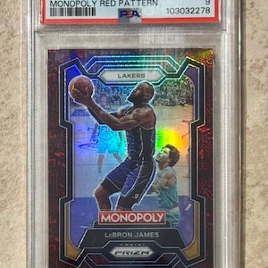 Puede incluir: Una tarjeta coleccionable de 2023 Prizm Monopoly LeBron James, calificada por PSA. La tarjeta presenta un patrón rojo y una imagen de LeBron James jugando al baloncesto. La tarjeta está en un estuche protector.
