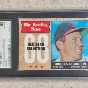 Könnte beinhalten: Eine Baseballkarte von Brooks Robinson von 1968 von Topps, bewertet mit 92 von SGC. Die Karte zeigt ein Porträt von Robinson mit Baseballkappe, mit dem Text "The Sporting News" und "All Star Selection". Die Karte ist in einem Schutzhalter eingeschlossen.