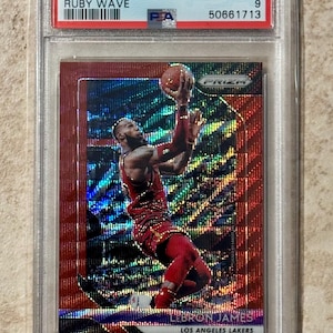 Può includere: Una carta da basket 2018 Panini Prizm LeBron James Ruby Wave, valutata Mint 9 da PSA, in un supporto protettivo. La carta mostra LeBron James in azione, con una divisa rossa dei Lakers. È visibile il numero 6.