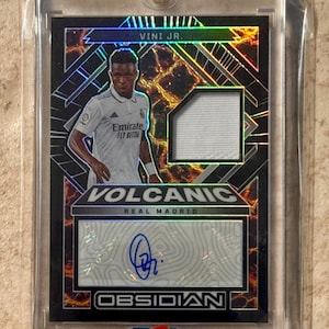 Pode incluir: Um cartão colecionável Vini Jr. Real Madrid Obsidian Volcanic em um protetor. O cartão apresenta uma imagem do jogador, um pedaço de camisa e um autógrafo. O cartão tem um design preto e laranja com o texto "OBSIDIAN".