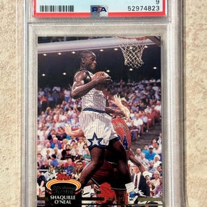 May include: A 1992 Stadium Club Shaquille O'Neal basketball card, graded PSA Mint 9, #247. The card features an action shot of the player in a white pinstripe uniform with a blue star, holding a basketball mid-air during a game in a crowded arena.