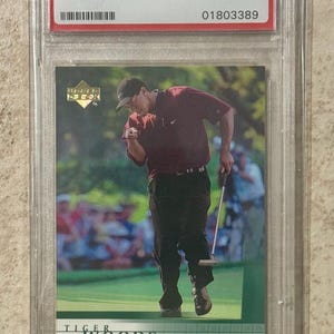 Puede incluir: Una tarjeta coleccionable de golf Upper Deck 2001 con Tiger Woods, calificada MINT 9 por PSA. La tarjeta muestra a Woods en acción en un campo de golf verde, con una camisa granate y pantalones negros. La tarjeta está en un protector transparente.