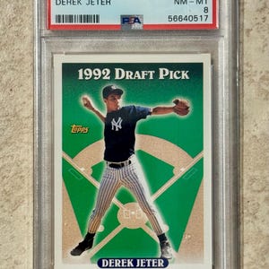 May include: A 1993 Topps Derek Jeter baseball card, graded NM-MT 8 by PSA, encased in a protective holder. The card features a 1992 Draft Pick image of Jeter in a New York Yankees uniform, set against a green and tan baseball field.