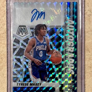 May include: A basketball trading card featuring Tyrese Maxey of the Philadelphia 76ers. The card has a mosaic design with the word "AUTOGRAPHS" and "ROOKIE" visible. Maxey is shown in his blue 76ers uniform, holding a basketball. The card is encased in a protective sleeve.