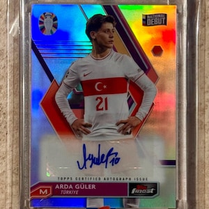 May include: A collectible trading card featuring Arda Güler in a white soccer jersey with the Turkish flag and the number 21. The card has a holographic design with the text "National Debut" and "Topps Certified Autograph Issue."